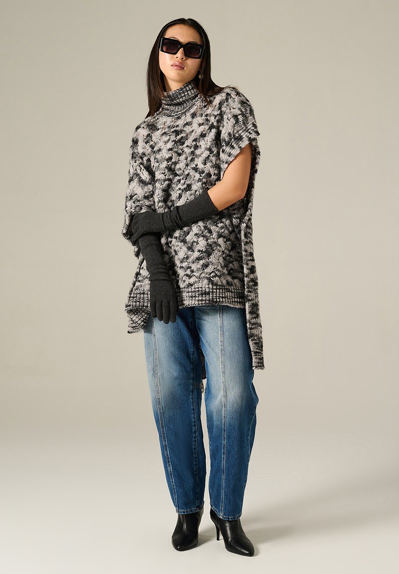 Poncho overdimensionato lavorato a maglia in grigio con un motivo texturizzato, indossato con guanti neri e stivali con tacco, abbinato a jeans blu larghi.