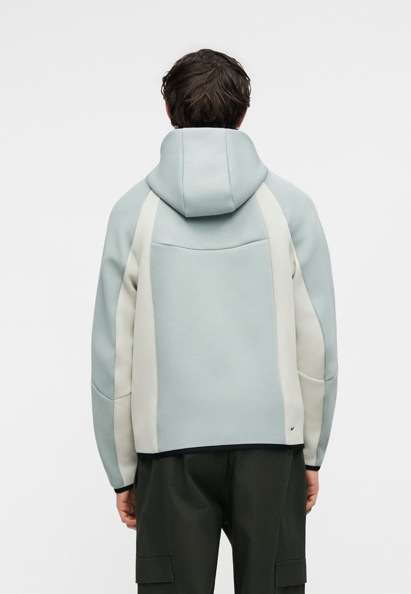 Mann trägt hellblauen und weißen Hoodie mit Kapuze, kombiniert mit dunklen Cargohosen, steht mit dem Rücken zur Kamera vor einfarbigem Hintergrund.