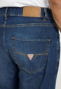 Jeans in denim blu con un'etichetta in pelle marrone con "GUESS 1981" e un logo triangolare rosso e bianco sulla tasca posteriore.