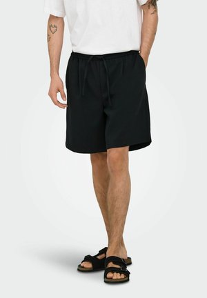 Hombre con pantalones cortos negros hasta la rodilla con cordón, camiseta blanca, sandalias negras y tatuajes visibles en los brazos, de pie frente a un fondo liso.