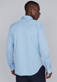 Chemise à manches longues bleu clair avec un col à boutons, ourlet courbé et poignets à un bouton. Texture lisse, le dos présente une boucle de suspension.