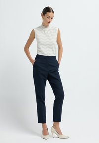 Valkoinen hihaton toppi, jossa on sahauskuvio, yhdistettynä räätälöityihin tummansinisiin raidallisiin housuihin ja beigeihin korkokenkiin. Minimalistinen muotoilu.