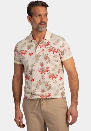 Man met korte baard die een beige polo met korte mouwen en bloemenprint draagt en lichtbruine broek draagt, kijkt opzij tegen een effen achtergrond.