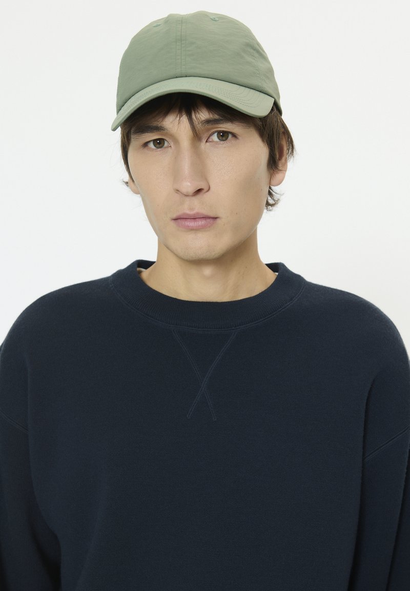 Casquette en coton verte à visière incurvée, associée à un sweat-shirt bleu marine avec un col rond et un détail de point de croix sur l'épaule.