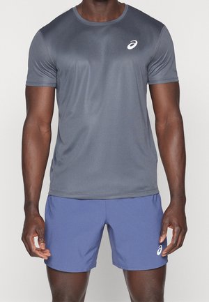 Atletische man draagt een strak grijs T-shirt met korte mouwen en blauwe short, beide met een wit Asics-logo aan de linkerkant.