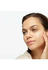 Crème hydratante appliquée sur la joue d'un modèle ; texture lisse et crémeuse, légère brillance. Le modèle a les cheveux châtain clair et des yeux verts, portant un haut beige.