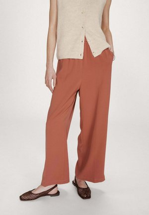 Femme portant un pantalon large couleur rouille, un haut sans manches beige boutonné et des ballerines à bride arrière en maille marron, debout avec une main dans la poche.