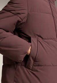 Bordo puffer jakna s kestenjastom teksturom, koja ima bočnu džep i opušteni kroj. Tkanina izgleda glatko i izolirano.