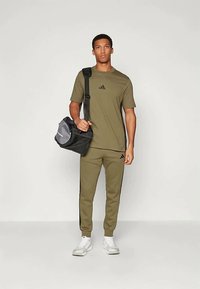 Oliivroheline spordirõivas mustade detailidega, sealhulgas lühikeste varrukatega särk ja kitsenevad püksid. Must kott hallidedetailidega. Valged spordijalatsid.