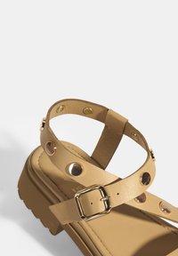 Beige Plattformsandalen aus Leder, mit einem dünnen Knöchelriemen, einer goldenen Schnalle und dekorativen goldenen Nieten entlang des Riemens.