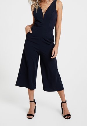 Kvinde iført en navyblå ærmeløs jumpsuit med vide ben og sorte høje hælede sandaler med ankelrem, stående med den ene hånd i lommen.