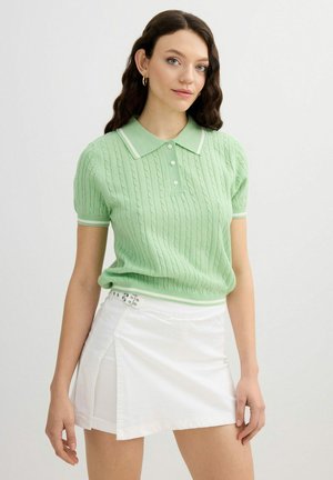 Joven con pelo ondulado lleva un polo de punto verde claro de manga corta y una minifalda blanca envolvente con detalle de hebilla.