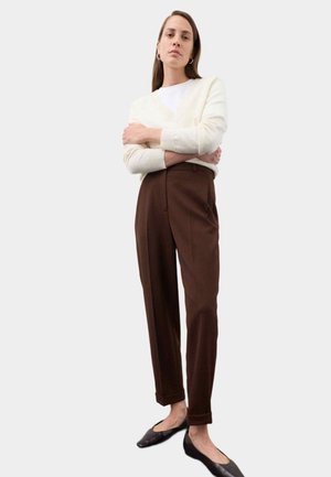 Kvinde stående med armene over kors iført hvid sweater, brune bukser med høj talje og sorte flade sko mod ensfarvet baggrund.