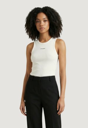 Mladá žena s kudrnatými vlasy, která má na sobě bílý bezrukávový žebrovaný top a černé kalhoty s vysokým pasem, stojí před jednobarevným pozadím.