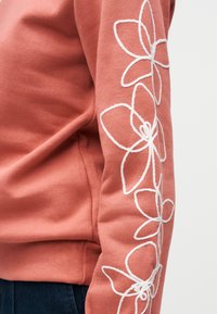 Μανίκι φούτερ σε σκουριά απόχρωση με λευκά κεντημένα λουλουδάτα σχέδια κατά μήκος του βραχίονα, φορεμένο με σκούρο μπλε παντελόνι.