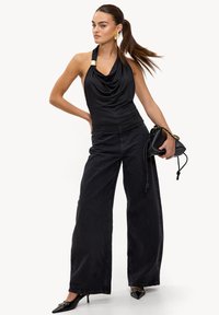 Haut noir à col cheminée avec un accent doré, associé à un jean noir à jambes larges. Le mannequin tient un petit sac à main noir et porte des escarpins noirs pointus.