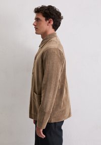 Corduroy jasje in lichtbruin met een overhemdkraag, knoopsluitingen, twee voorzakken en een gestructureerde afwerking. Gemaakt met een donkere broek.