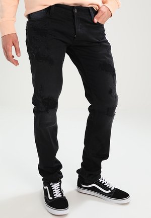Jean en denim noir avec des patchs usés, coupe slim, design à cinq poches, et surpiqûres contrastantes. Porté avec des baskets noires et blanches.