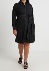 Černé denimo šaty košile s dlouhými rukávy, zapínání na knoflíky, boční kapsou, stahováním v pase a kontrastním prošíváním. Vytvořeno s černými sandály.