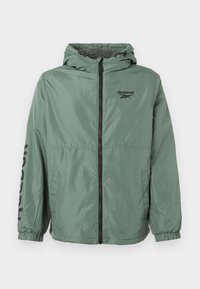 Žalia lengva striukė su kapišonu, kuriuose yra juodas užtrauktukas ir didelis "Reebok" logotipas ant rankovės. Elastingi rankogaliai ir dvi šoninės kišenės.