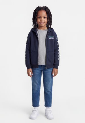 Παιδί με κοτσίδες, φοράει μπλε σκούρο φούτερ με φερμουάρ, γκρι μπλουζάκι, μπλε τζιν και άσπρα αθλητικά παπούτσια, στέκεται μπροστά σε απλό φόντο.