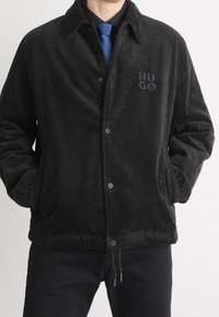 Homme portant une veste en velours côtelé noire avec le logo "HUGO" sur la poitrine, une cravate bleue et une chemise noire, les mains dans les poches, sur un fond clair uni.
