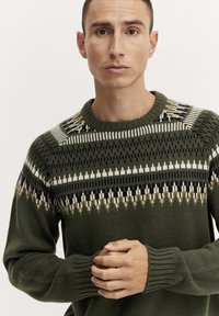 Grüner gestrickter Pullover mit geometrischen Mustern in Weiß, Gold und Schwarz. Gerippte Bündchen und runder Halsausschnitt. Strukturiertes, warmes Material.