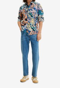 Kleurrijk overhemd met lange mouwen en knopen, voorzien van abstracte patronen in blauw, oranje en groen, gecombineerd met lichtblauwe jeans en crèmekleurige sneakers.
