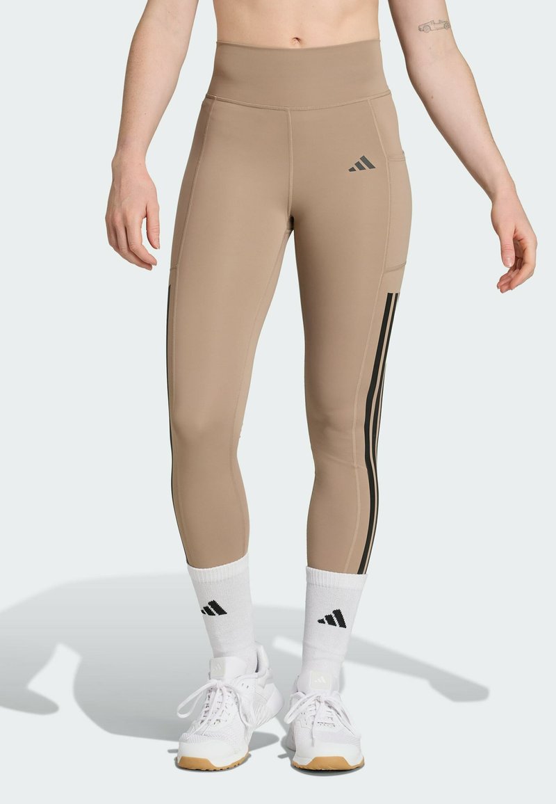 Beige high-waisted leggings with black side stripes and textured fabric. Paired with white shoes and socks featuring the Adidas logo.