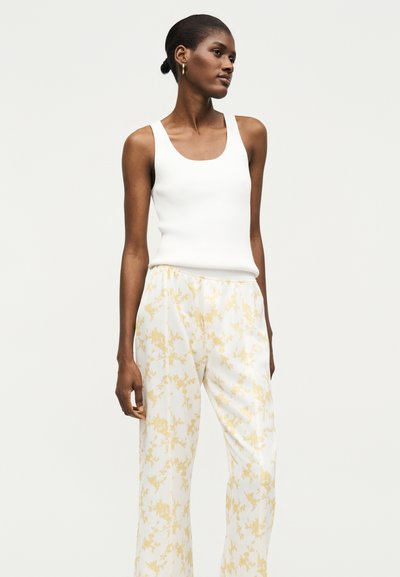 Γυναίκα με σκουρόχρωμο δέρμα και μαλλιά σε κότσο φοράει λευκό αμάνικο τοπ και φαρδιά λευκά παντελόνια με ανοιχτά κίτρινο λουλουδάτο μοτίβο, στέκεται μπροστά σε απλό φόντο.