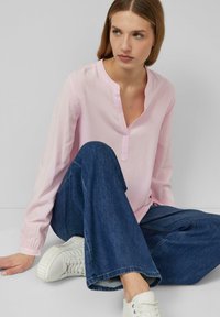 Jasnoróżowa bluzka z długim rękawem i dekoltem w szpic, zestawiona z ciemnoniebieskimi jeansami o szerokich nogawkach i białymi trampkami, siedząca na tle w stonowanych kolorach.