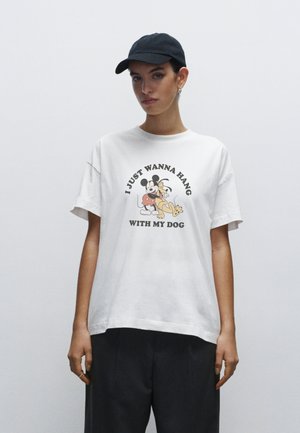 Hvid bomulds t-shirt med korte ærmer, der har et cartoonprint af en hund og karakterer. Casual pasform, suppleret med mørke shorts og en sort kasket.