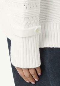 Witte gebreide trui met geribde manchetten, een knoopsluiting en een textuurpatroon langs het lichaam. Hand rustend op een donkere ondergrond.
