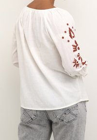 Blouse en coton blanc à manches longues, ornée de motifs floraux brodés en rouge sur les bras. Texture douce et coupe décontractée, col rond.
