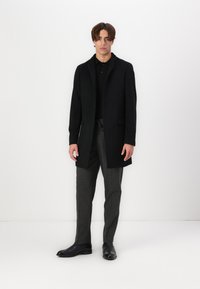 Cappotto nero con colletto, chiusura a un bottone e design strutturato. Indossato sopra un polo nero e pantaloni grigi a righe. Stivali neri.