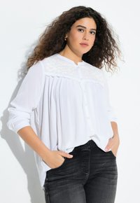 Donna con capelli ricci che indossa una camicia bianca a bottoni testurizzata e jeans neri, in piedi con le mani in tasca su uno sfondo bianco.