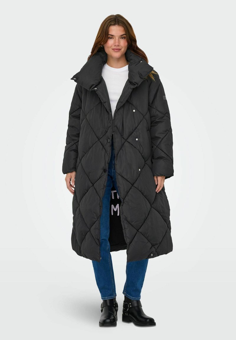 Ilgas juodas pūkinis paltas su deimantiniu siuvinėtu raštu, aukštas apykaklė, spaudimo sagos, dėvimas virš baltos marškinių ir mėlynų džinsų, juodi kulkšnies auliniai batai.