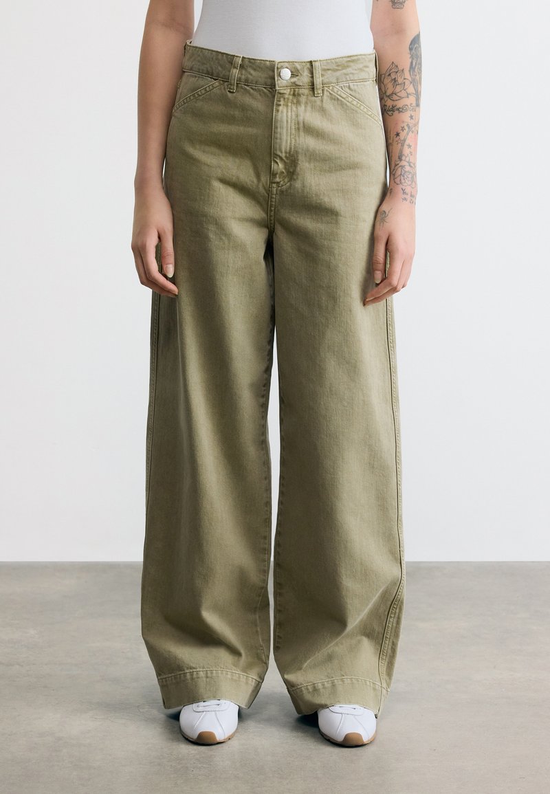 Άτομο που φορά άνετο παντελόνι χακί και λευκά αθλητικά παπούτσια, στέκεται σε γκρι πάτωμα με λευκό φόντο.