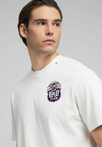 Vit bomulls-t-shirt med en cirkulär broderad logotyp föreställande en örn och texten "REPLAY TEAM" i blått över bröstet. Kortärmad med rund halsringning.