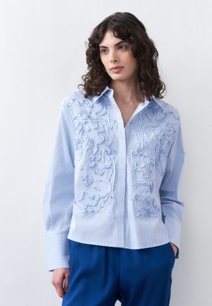 Γυναίκα με καστανά σγουρά μαλλιά φοράει μπλούζα με ριγέ μπλε και λευκό σχέδιο και τρισδιάστατη κεντητή λουλουδιαστή διακόσμηση, συνδυασμένη με μπλε παντελόνι, σε απλό φόντο.