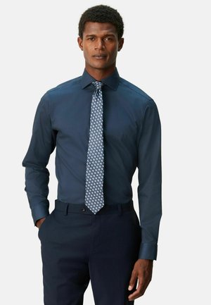 Man wearing a long-sleeve dark blue dress shirt and patterned blue tie, with dark blue pants, standing with one hand in pocket.