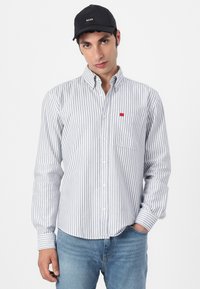 Rutet skjorte med knapper i lyseblå og hvit, med brystlomme og liten rød etikett. Sammen med lyseblå jeans og en svart caps.