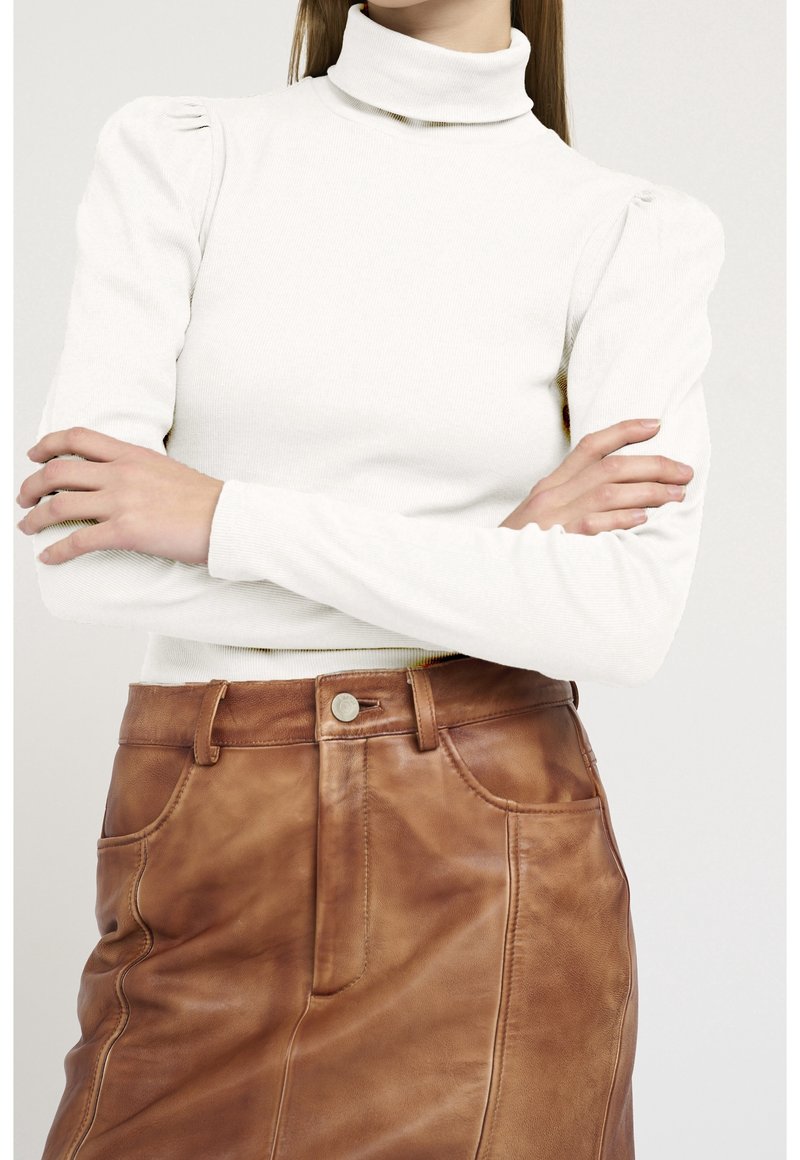 Femme bras croisés portant un col roulé blanc à manches bouffantes et une jupe en cuir marron taille haute.