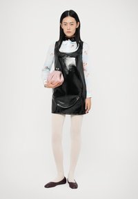 Γυναίκα φορώντας γυαλιστερό μαύρο αμάνικο φόρεμα πάνω από εμπριμέ μπλούζα, λευκές κολάν, μπορντό μπαλαρίνες, κρατώντας μικρή ροζ τσάντα σε απλό φόντο.