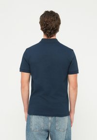 Άνδρας με κοντά καστανά μαλλιά, ντυμένος με μπλε ναυτικό πόλο με κοντά μανίκια και ανοιχτόχρωμο τζιν, απεικονισμένος από πίσω.