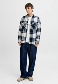 Joven de pie, vistiendo una camisa de cuadros azul marino y blanca sobre un henley blanco, jeans oscuros anchos y zapatos beige sobre un fondo blanco.