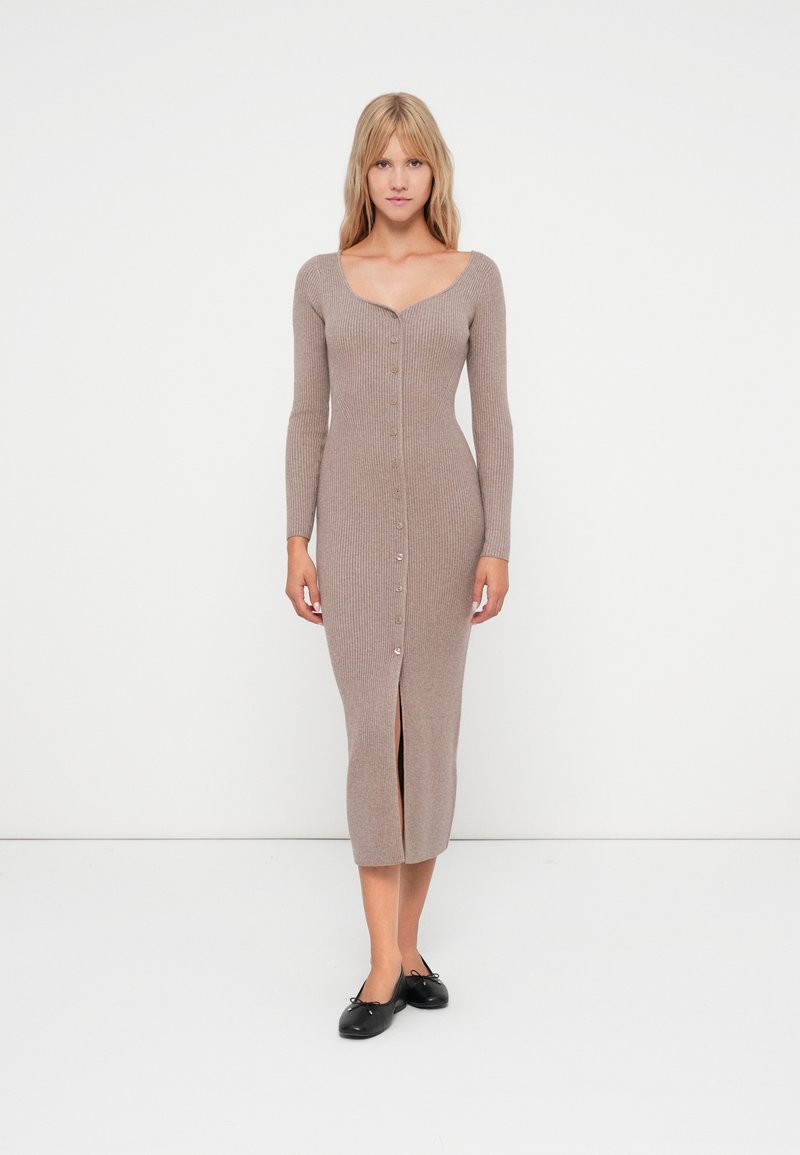 Μακριά, ριμπ πλεκτή φόρμα σε ανοιχτό καφέ χρώμα με V λαιμόκοψη, μακριά μανίκια, κουμπιά μπροστά και σχισμή στο κάτω μέρος. Φοριέται με μαύρες μπαλαρίνες.