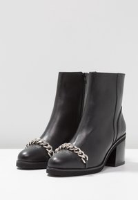 Bottes basses en cuir noir avec un bout arrondi, ornées d'une chaîne argentée sur le devant et dotées d'un talon carré. Fermeture éclair latérale.