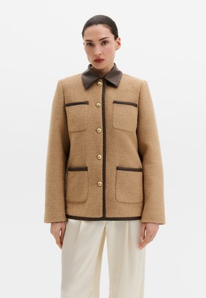 Vrouw draagt een beige gestructureerd jasje met donkerbruine bies en gouden knopen, gecombineerd met losse witte broek, tegen een effen achtergrond.