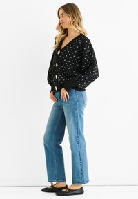 Black knitted cardigan with white polka dots, v-neck, oversized sleeves, front buttons, paired with high-waisted blue jeans and black flat shoes.